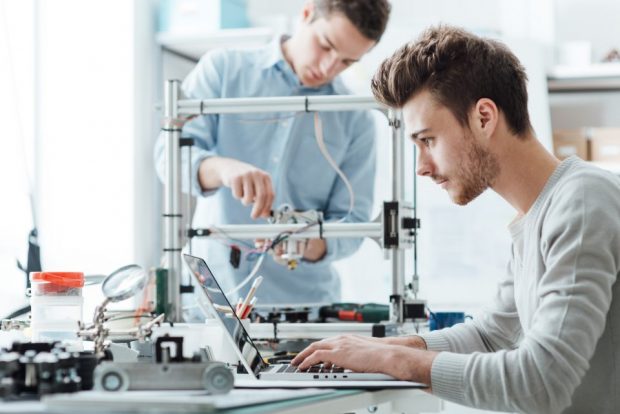 A man works a laptop while another uses a tool to fine tune an electronic circuit. 