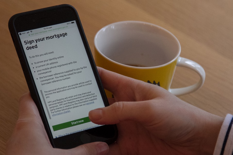 A hand holds a mobile phone showing the Sign your mortgage deed service on its screen, with a coffee mug on the desk alongside.