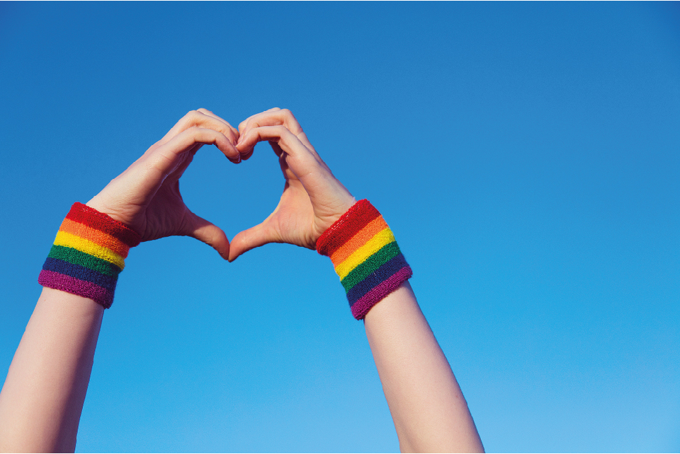 Two hands forming a heart shape with rainbow wristbands around the wrists.