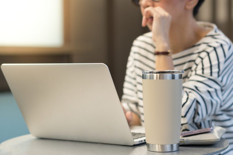 An office worker using a laptop with a reusable stainless steel tumbler mug alongside.