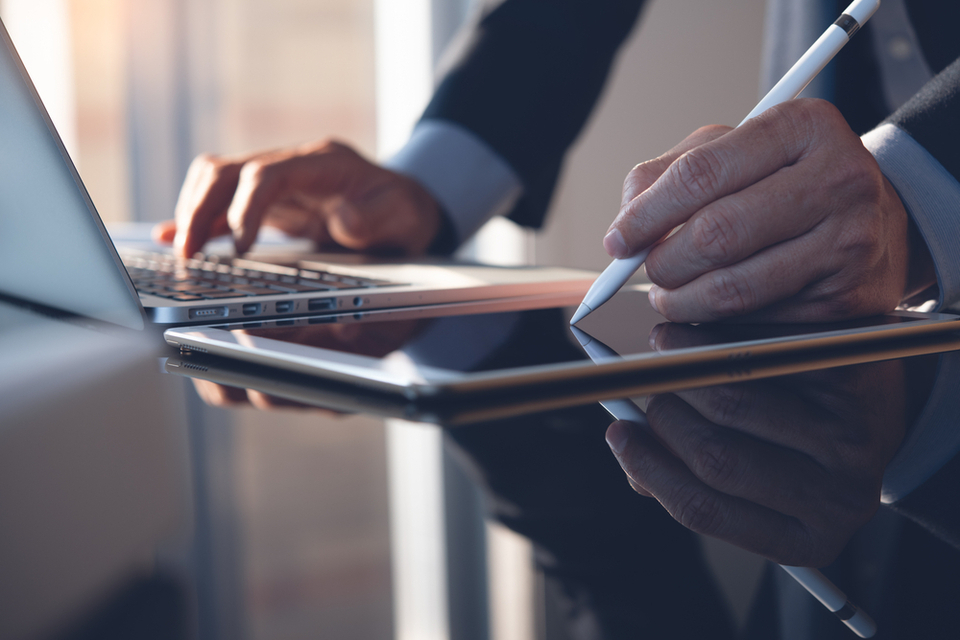 Man in suit using stylus pen to sign contract on digital tablet while working on laptop computer.