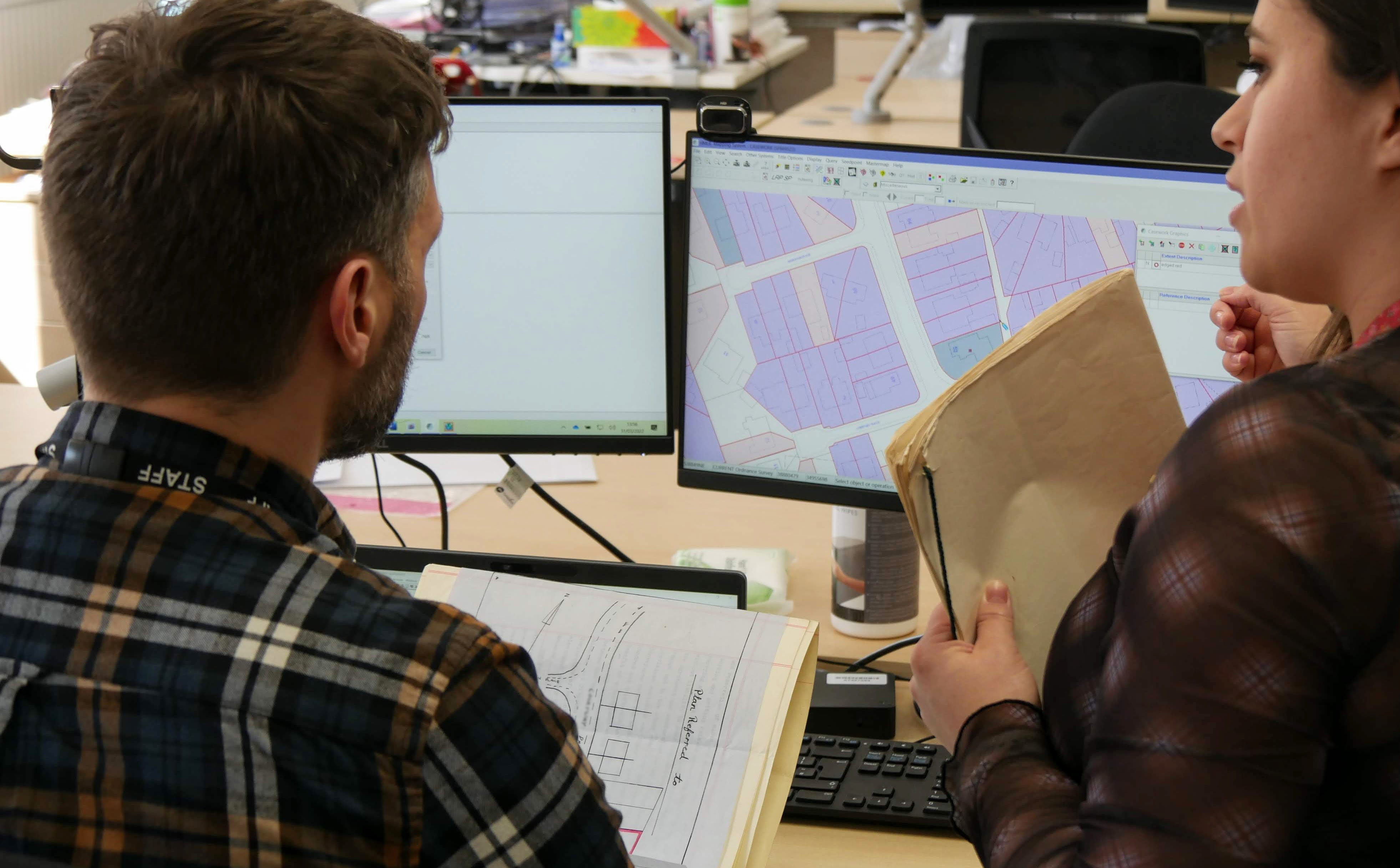 On the left, a male caseworker in a plaid shirt looks at a mapping screen on his computer, while a female caseworker to his right offers advice.