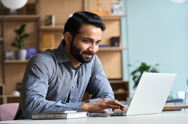 A smiling man with a beard sitting at a desk with a laptop open in front of him.