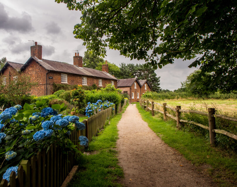 A footpath runs between fences, with a field and overhanging tree beyond the right-hand fence and a garden and row of cottages beyond the left.