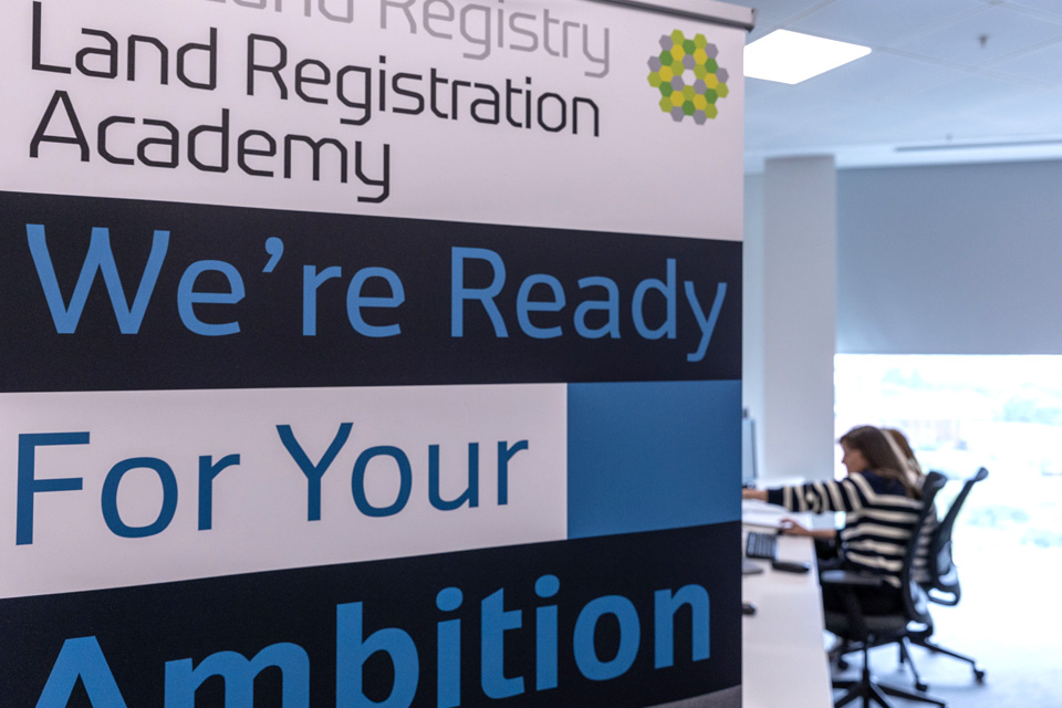 A banner saying 'Land Registration Academy' and 'We're ready for your ambition', with caseworkers sitting at a desk behind.